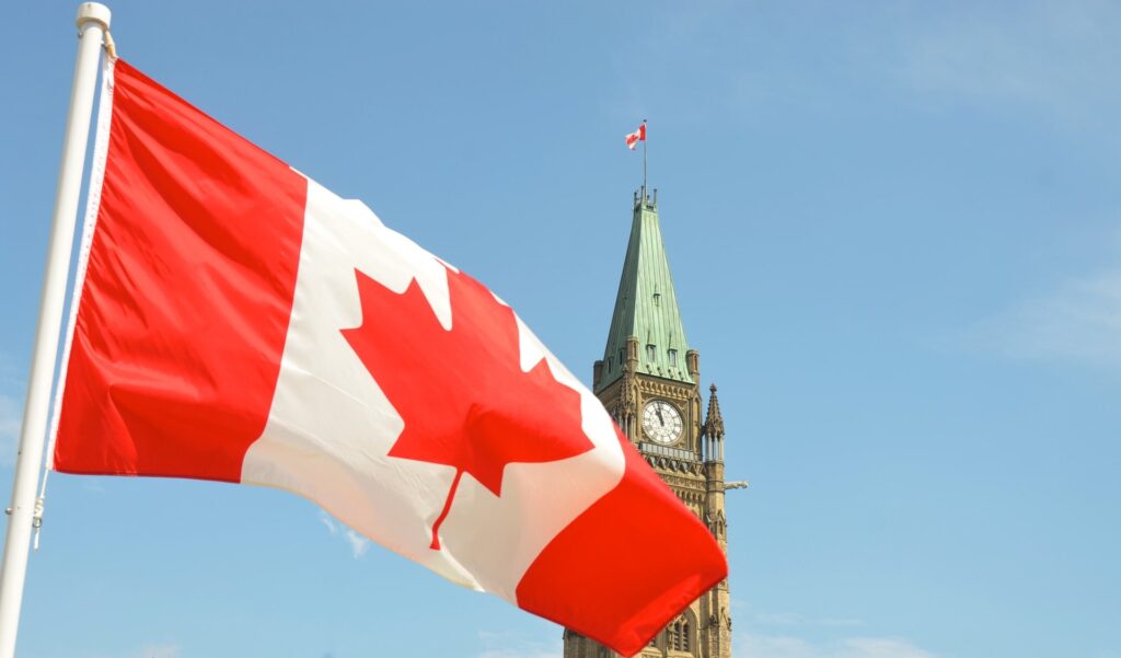 A photo of the Canadian flag being blown by the wind. A tall infrastructure with a large clock near its roof can also be seen in the background. Ths is the National Gallery of canada, one of the best museums in Ottawa.