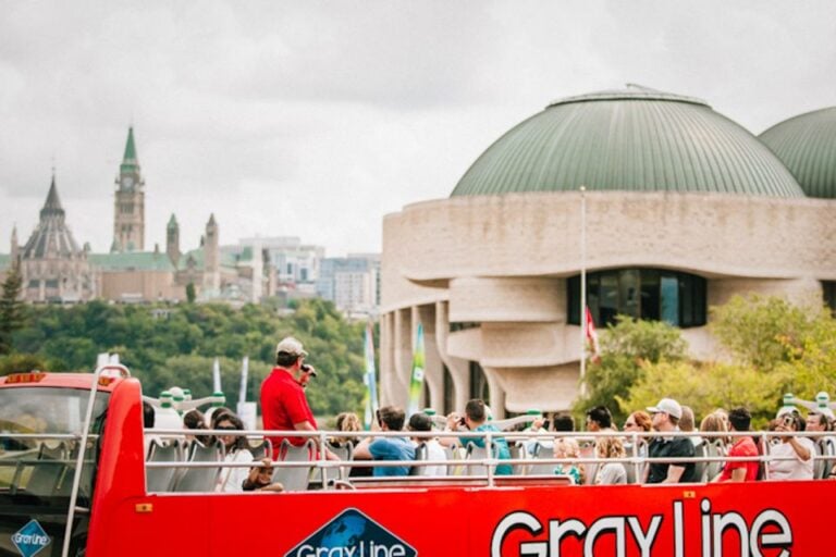 "Grayline" tour bus that is a double decker. It's bright red as it passes by the Museum of Nature in Ottawa, with a view of Parliament Hill across the Ottawa River in the distance. A man is leading the hop on hop off tour for visitors on the top level of the bus.